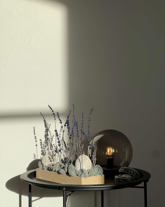 Composition florale avec un support en bois massif posé sur une table de nuit noir avec une lumière en verre. Le lichen stabilisé est bleu et avec de la lavande et autre feuillage.