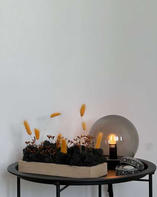 Composition florale avec un support en bois massif posé sur une table de nuit noir avec une lumière en verre. Le lichen stabilisé est noir et est composé de chouchou couleur abricot et limonium stabilisé orange.