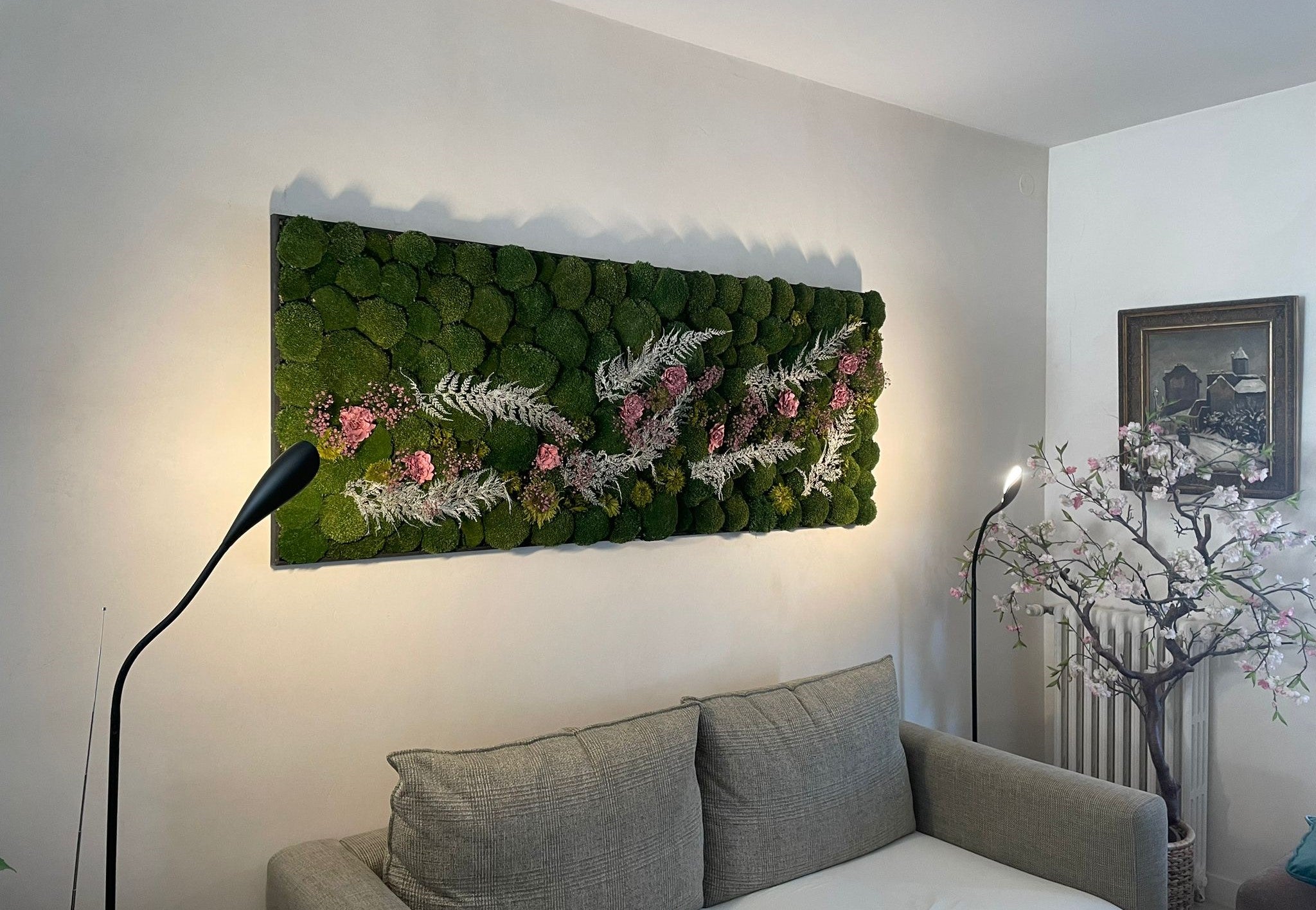 Tableau en végétaux stabilisé dans un salon modern avec un canapé gris et deux lumières design. Le tableau est composé de mousse verte naturel et de fleurs colorés et de fougères blanches.