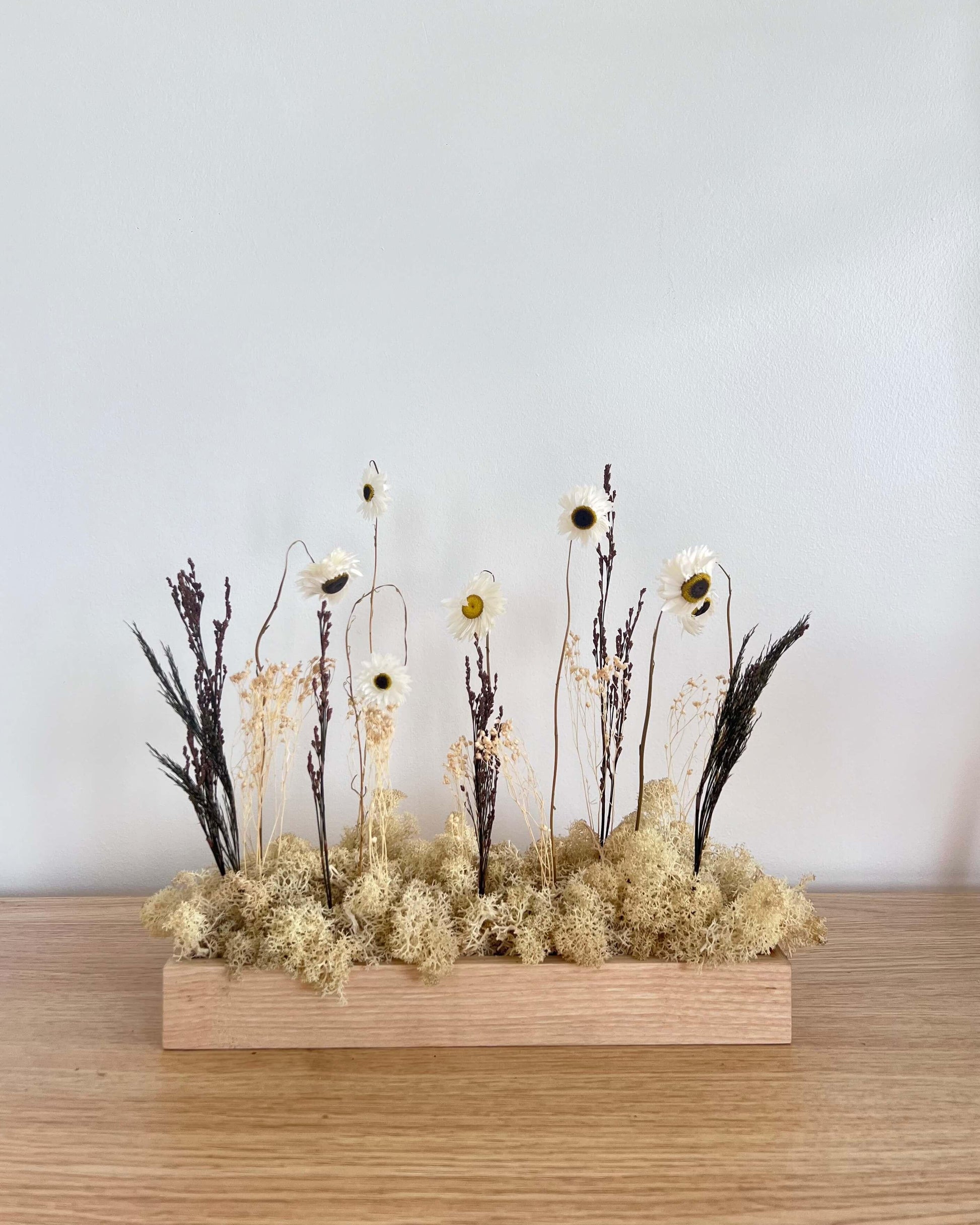 Composition florale avec un support en bois massif posé sur une tablette en bois. Le lichen stabilisé est naturel et est composé de fleurs et autre feuillage.