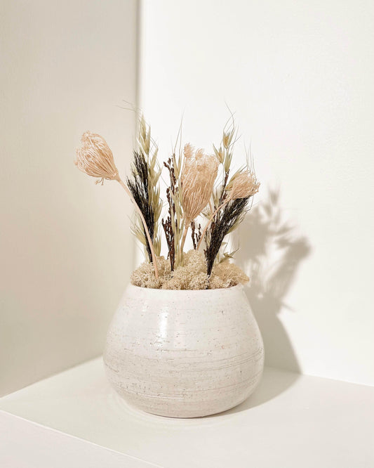 Composition florale moderne dans une poterie blanche posé sur une tablette blanche. Composition avec du lichen stabilisés naturel et des plantes dans les tons naturels.