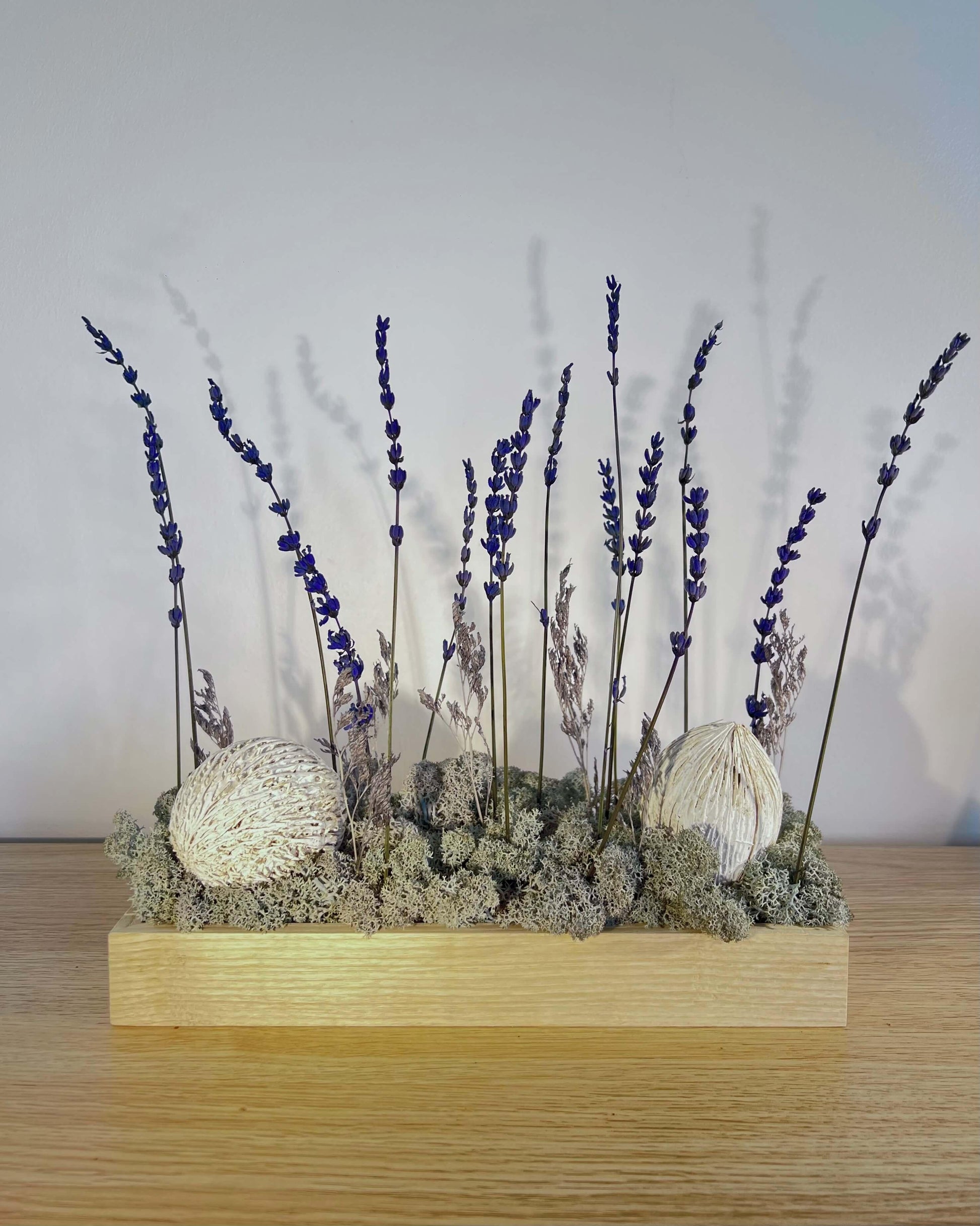 Composition florale avec un support en bois massif posé sur une tablette en bois massif. Le lichen stabilisé est bleu et avec de la lavande et autre feuillage.