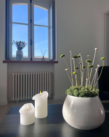Composition florale moderne dans une poterie blanche posé sur une table noir avec deux bougies blanches allumés. Composition avec du lichen stabilisés vert forêt et des plantes vertes et blanches.