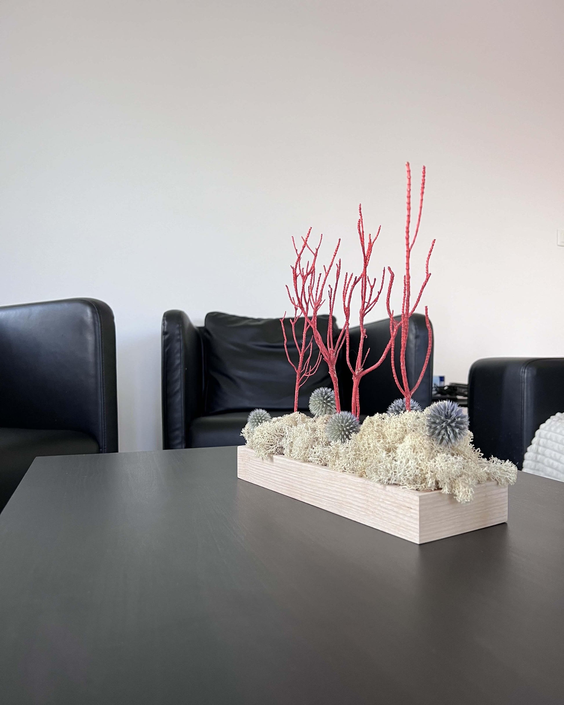 Composition florale avec un support en bois massif posé sur une table noir de salon moderne. Le lichen stabilisé est naturel et est composé de chardon bleu et de romarin orange/rouge.