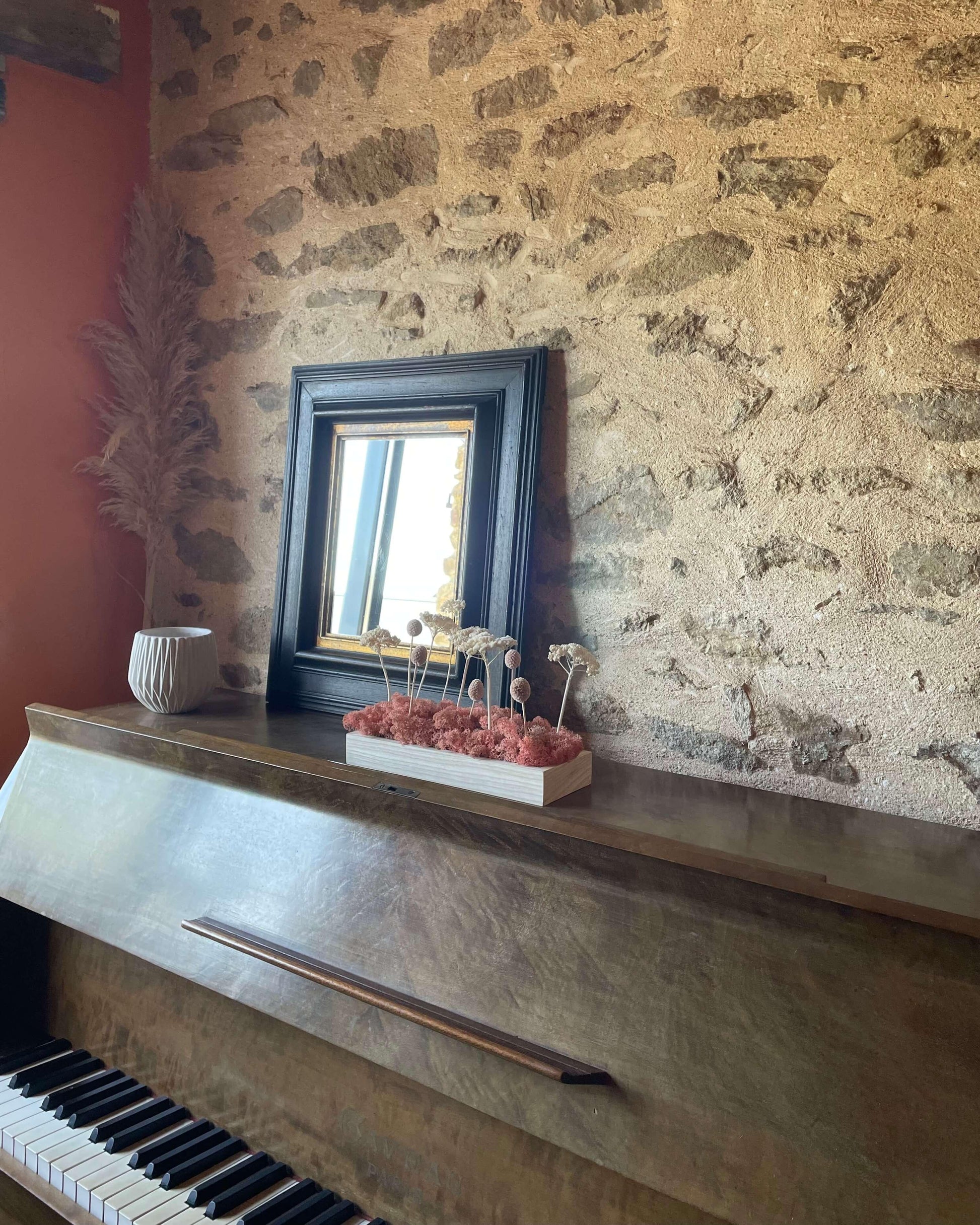 Décoration intérieure au design floral en lichen stabilisé et plantes naturelles stabilisées dans une maison contemporaine. Composition en végétaux stabilisés posé sur un piano devant un miroir, et un mur en pierre. La composition est sur les tons roses..