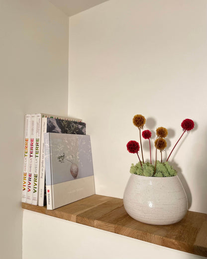 Composition florale moderne dans une poterie blanche posé sur une tablette en bois massif à coté de livres. Composition avec du lichen stabilisés vert claires et des fleurs modernes jaune te rouge.