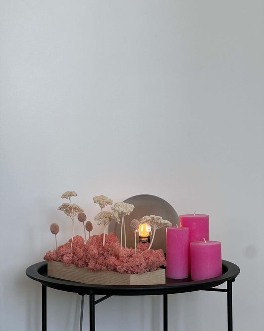 Composition florale avec un support en bois massif posé sur une table de nuit noir avec des bougie roses et une lumière en verre. Le lichen stabilisé est rose et est composé de achillée blanche et craspédie rose.