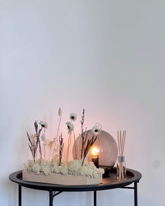Composition florale avec un support en bois massif posé sur une table de nuit noir à coté d'une lumière en verre et d'un sens bon. Le lichen stabilisé est naturel et est composé de fleurs et autre feuillage.