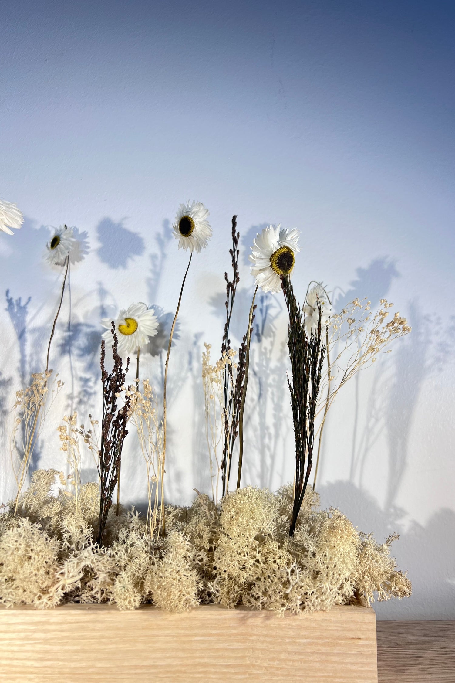 Zoom sur composition florale avec un support en bois massif posé sur une tablette en bois. Le lichen stabilisé est naturel et est composé de fleurs et autre feuillage.
